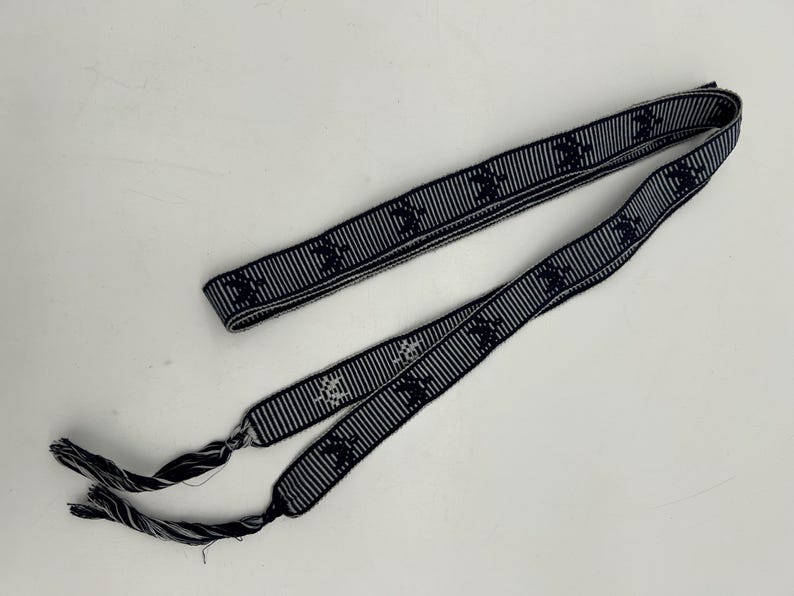 May include: A navy blue and white striped ribbon with a repeating pattern of a stylized emblem. The ribbon is folded and has frayed ends. The ribbon is lying on a white surface.