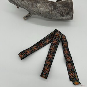 May include: Black ribbon with a floral pattern in orange, teal, and yellow. The ribbon is displayed against a white background, with a piece of driftwood in the upper portion of the image.