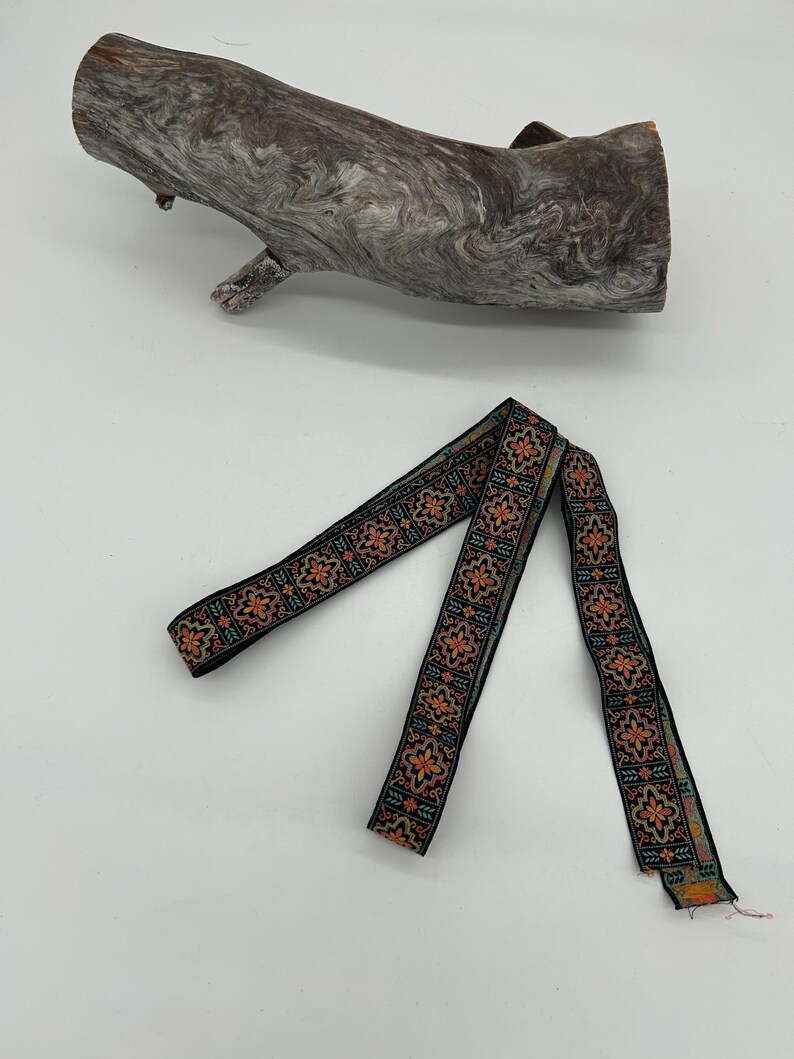 May include: Black ribbon with a floral pattern in orange, teal, and yellow. The ribbon is displayed against a white background, with a piece of driftwood in the upper portion of the image.