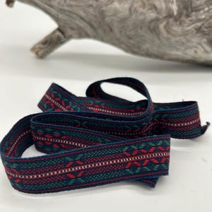 May include: Woven ribbon featuring a dark blue base with a repeating pattern of red, green, and white geometric shapes. The ribbon is curled on a white surface, with driftwood in the background. The ribbon's width is approximately 2 cm.