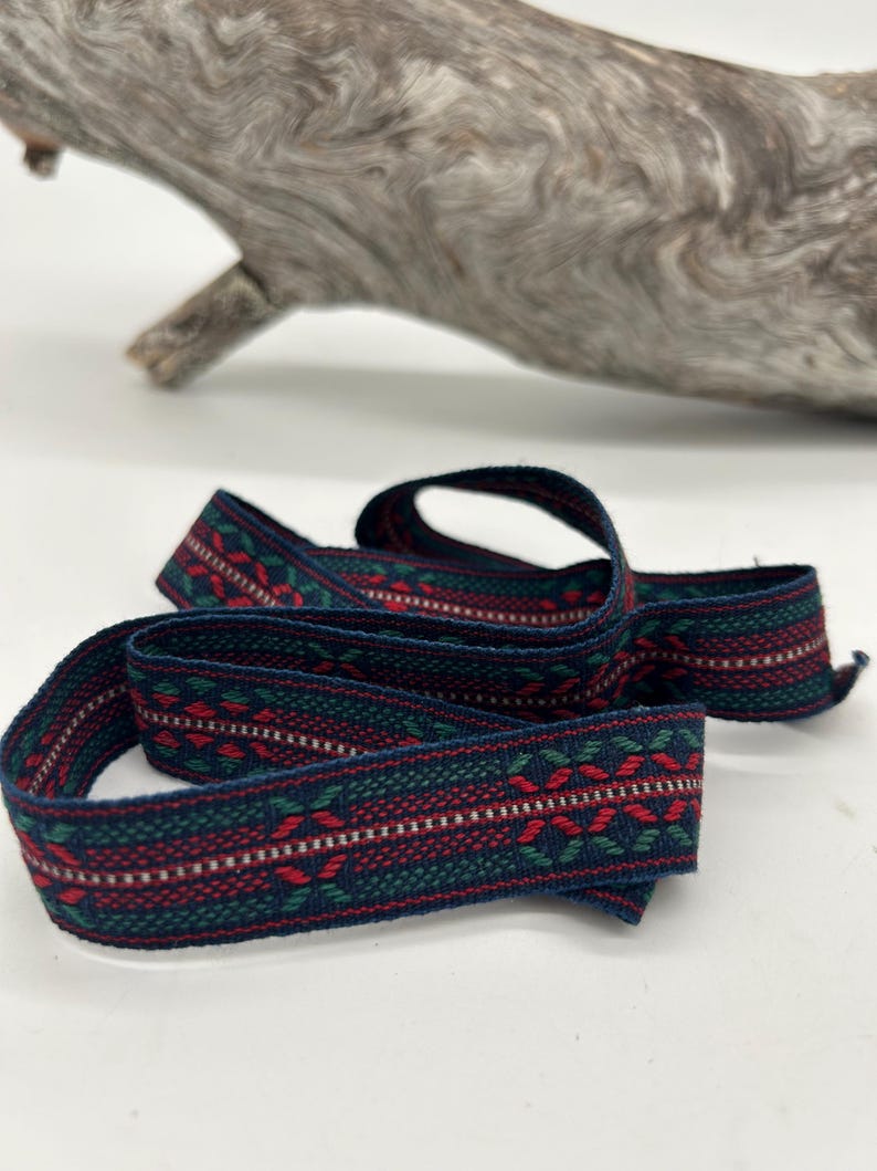 May include: Woven ribbon featuring a dark blue base with a repeating pattern of red, green, and white geometric shapes. The ribbon is curled on a white surface, with driftwood in the background. The ribbon's width is approximately 2 cm.