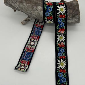 May include: Black ribbon featuring embroidered floral patterns in red, blue, and white, draped over driftwood. The floral design includes edelweiss flowers and other colorful blossoms. This decorative ribbon is suitable for crafts and embellishments.