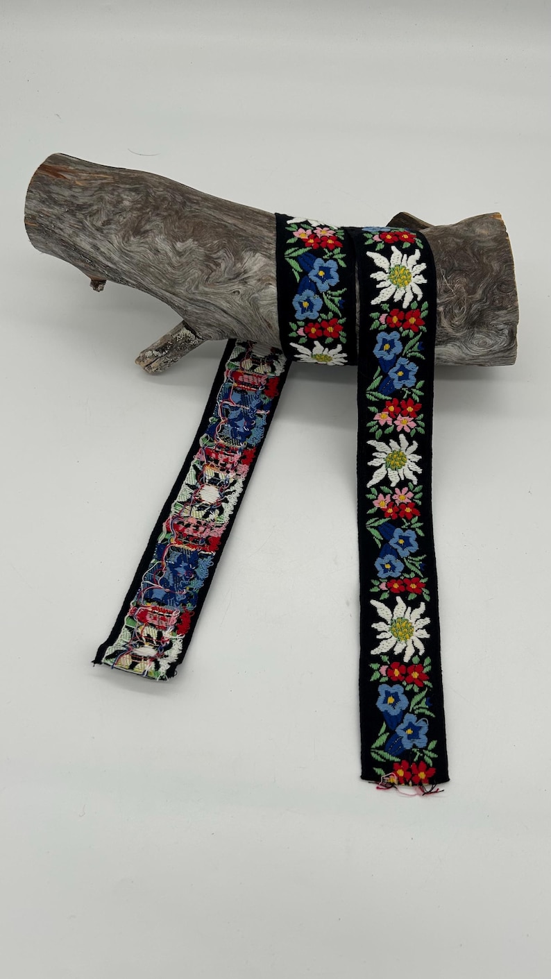 May include: Black ribbon featuring embroidered floral patterns in red, blue, and white, draped over driftwood. The floral design includes edelweiss flowers and other colorful blossoms. This decorative ribbon is suitable for crafts and embellishments.
