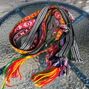May include: A long, woven fabric belt featuring intricate geometric patterns in vibrant red, yellow, green, black, and white. The textile band is coiled and finished with long, multi-colored yarn tassels at the ends, showcasing a dense, textured weave.