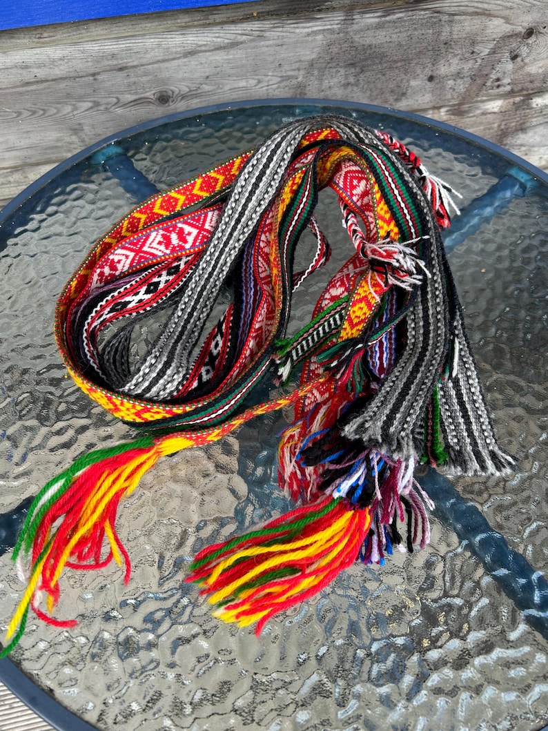 May include: A long, woven fabric belt featuring intricate geometric patterns in vibrant red, yellow, green, black, and white. The textile band is coiled and finished with long, multi-colored yarn tassels at the ends, showcasing a dense, textured weave.