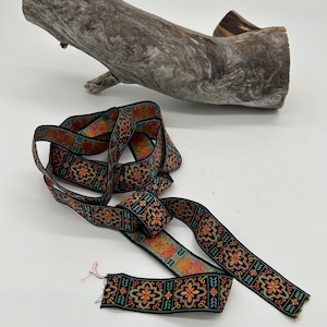May include: A decorative ribbon with a black background and a repeating floral pattern in orange, yellow, and teal. The ribbon is lying on a white surface next to a piece of driftwood.
