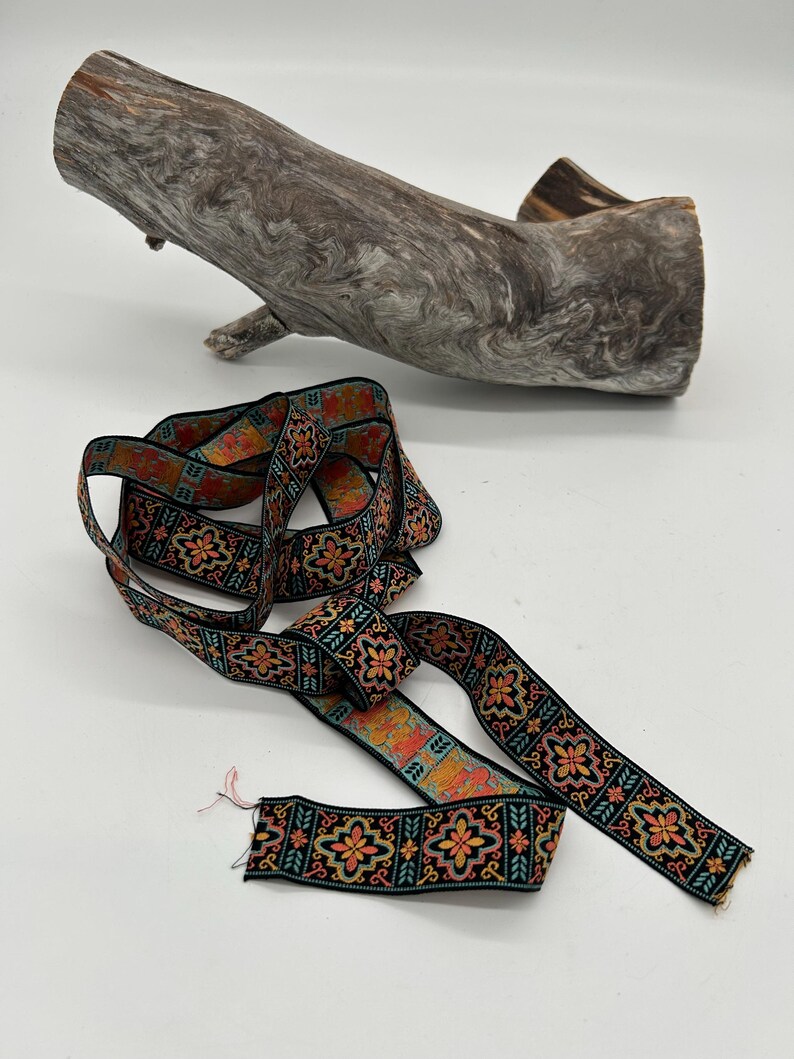 May include: A decorative ribbon with a black background and a repeating floral pattern in orange, yellow, and teal. The ribbon is lying on a white surface next to a piece of driftwood.