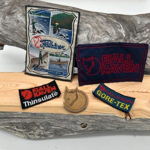 Puede incluir: Una colección de parches y un emblema de madera con forma de zorro. Los parches presentan el logotipo y el texto de Fjällräven, con un parche que muestra una ilustración escénica. Los parches son de varios colores, incluyendo azul, rojo y amarillo.