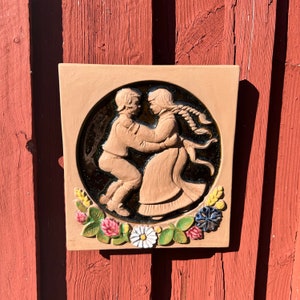 Puede incluir: Azulejo cerámico cuadrado, color beige, con un relieve de dos figuras bailando en un círculo. Las figuras visten ropa marrón claro. El círculo está rodeado por un borde floral con flores rosas, blancas y azules.