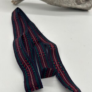 May include: Woven ribbon featuring a repeating pattern in red, green, and white on a dark blue background. The ribbon has a textured appearance and is curled. It is displayed on a white surface.