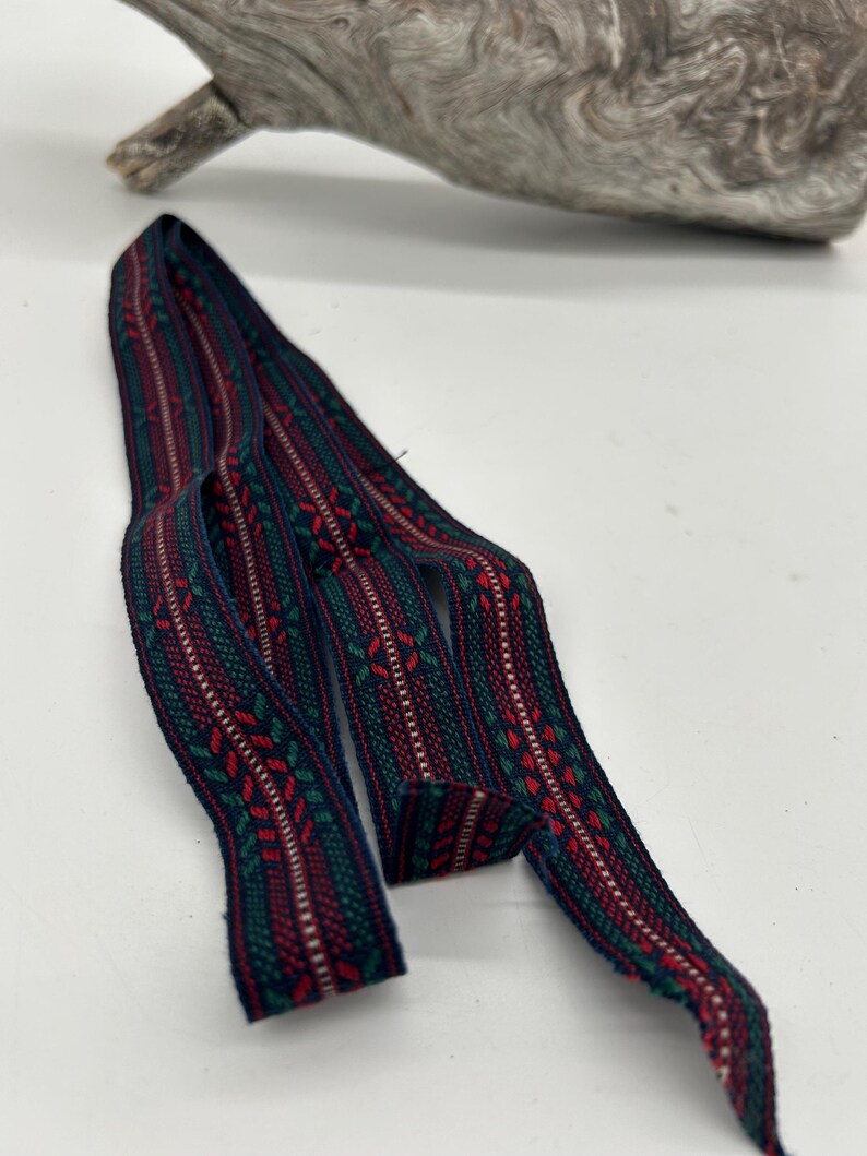 May include: Woven ribbon featuring a repeating pattern in red, green, and white on a dark blue background. The ribbon has a textured appearance and is curled. It is displayed on a white surface.