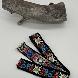 May include: Two black fabric straps with floral embroidery are shown. The embroidery features white, red, and blue flowers. A piece of driftwood is in the background.