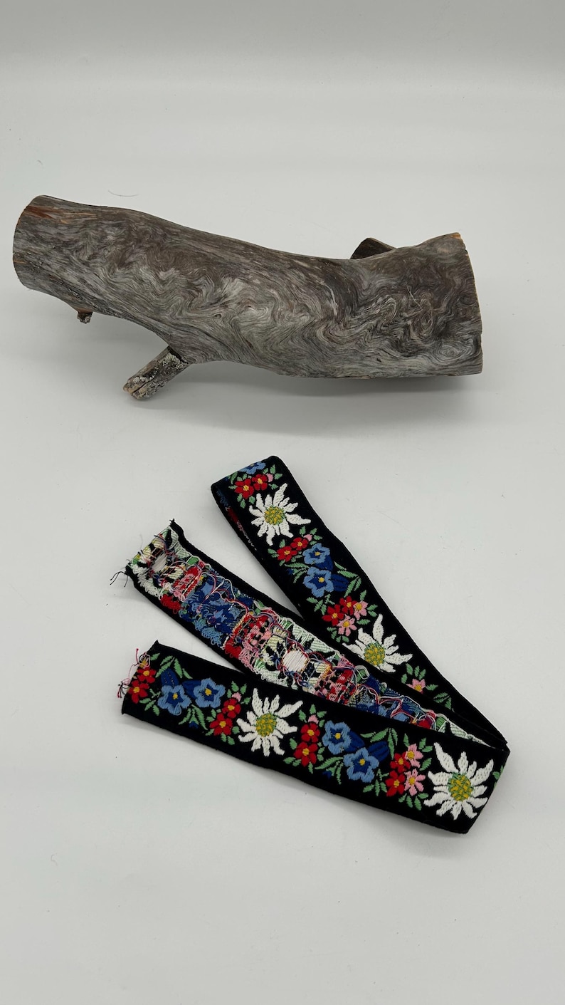 May include: Two black fabric straps with floral embroidery are shown. The embroidery features white, red, and blue flowers. A piece of driftwood is in the background.