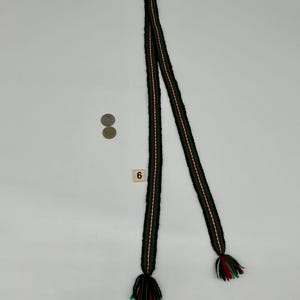 May include: A long, narrow woven belt or sash featuring a repeating pattern of black, green, and white stripes. The ends of the belt are finished with multi-colored yarn tassels in black, green, and white. A small square tile with the number 6 is present.