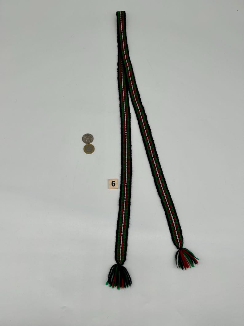 May include: A long, narrow woven belt or sash featuring a repeating pattern of black, green, and white stripes. The ends of the belt are finished with multi-colored yarn tassels in black, green, and white. A small square tile with the number 6 is present.