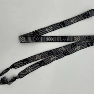 May include: A black and white woven belt with a geometric pattern. The belt has a repeating square design and tassels at the ends. The belt is made of a woven material and is likely used as an accessory.