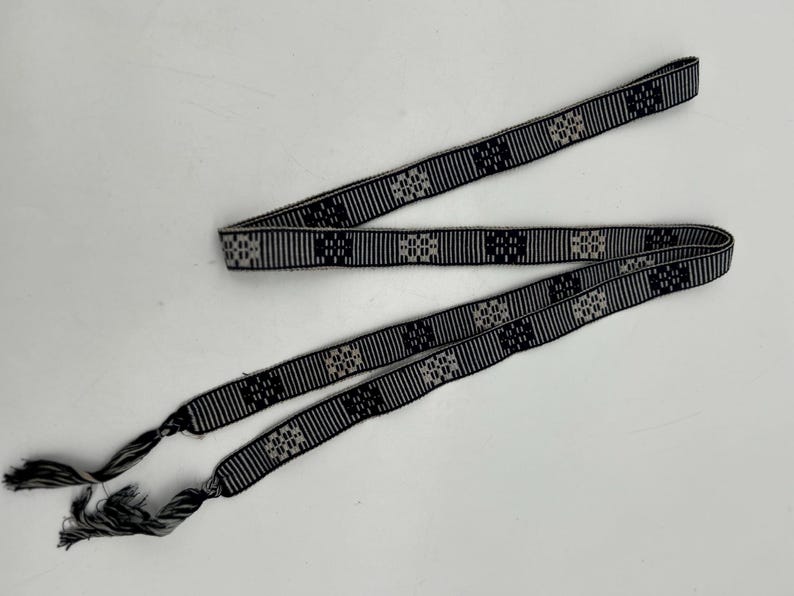 May include: A black and white woven belt with a geometric pattern. The belt has a repeating square design and tassels at the ends. The belt is made of a woven material and is likely used as an accessory.