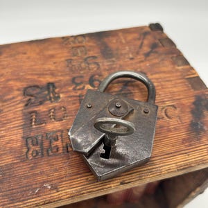 May include: A vintage metal padlock with a key inserted, resting on a wooden crate. The padlock is dark gray with a keyhole and a curved handle. The wooden crate has visible wood grain and stamped markings.