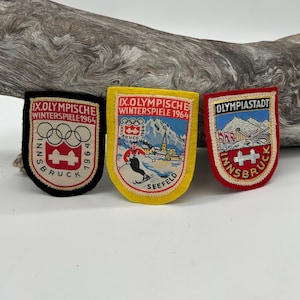 May include: Three vintage Olympic patches from the 1964 Winter Games in Innsbruck, Austria. The patches are in black, yellow, and red, with text and graphics related to the event. The patches feature the Olympic rings, the city name, and the year.