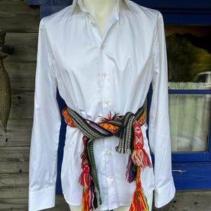 May include: A crisp white long-sleeved button-down shirt cinched at the waist with a woven sash. The sash features a complex geometric pattern in red, green, black, and yellow, finished with long, multi-colored yarn tassels hanging at the ends.