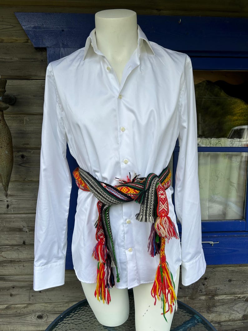 May include: A crisp white long-sleeved button-down shirt cinched at the waist with a woven sash. The sash features a complex geometric pattern in red, green, black, and yellow, finished with long, multi-colored yarn tassels hanging at the ends.