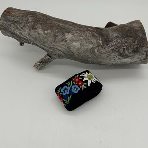 May include: A folded black ribbon with embroidered flowers in red, blue, and white. A piece of driftwood is in the background. The ribbon is likely used for crafts or embellishments.