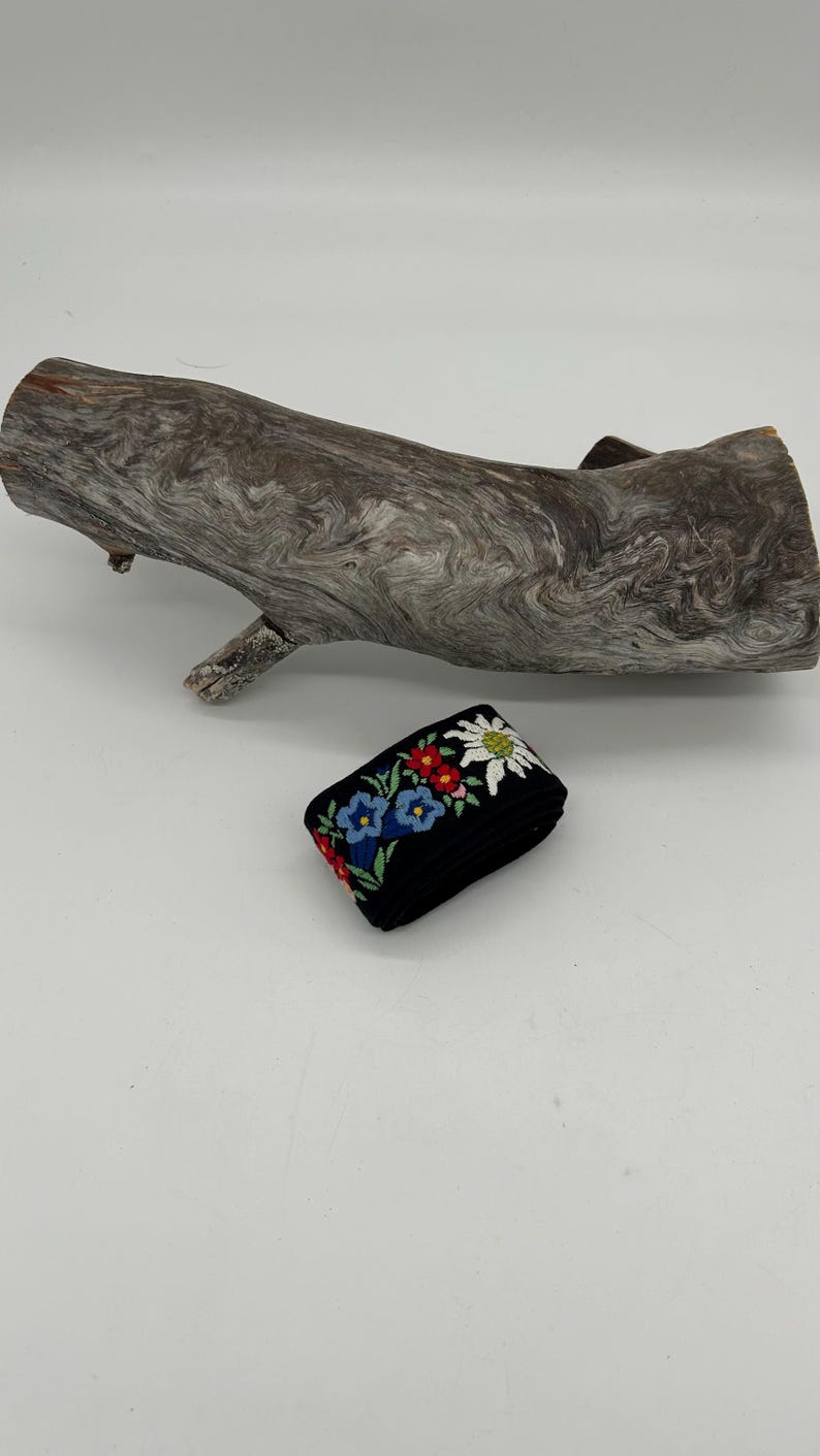 May include: A folded black ribbon with embroidered flowers in red, blue, and white. A piece of driftwood is in the background. The ribbon is likely used for crafts or embellishments.