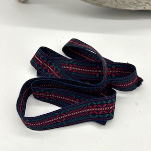 May include: Woven ribbon featuring a repeating pattern of red, green, and white geometric shapes on a navy blue background. The ribbon is folded and curved, displaying the detailed design. Suitable for craft projects, this ribbon is decorative.