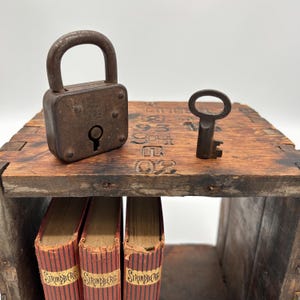 Puede incluir: Una estantería de madera antigua con aspecto envejecido, con un candado y una llave oxidados. Debajo se ven tres libros con cubiertas a rayas rojas y blancas. La superficie de la estantería tiene números grabados.