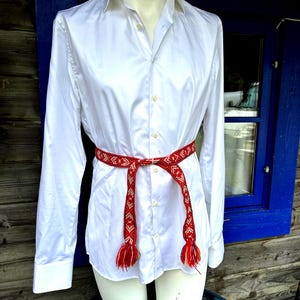 May include: A crisp white long-sleeved button-down shirt featuring a pointed collar and a central button placket. A narrow, woven belt with a red, white, and orange geometric pattern is tied around the waist, ending in two fringed tassels.