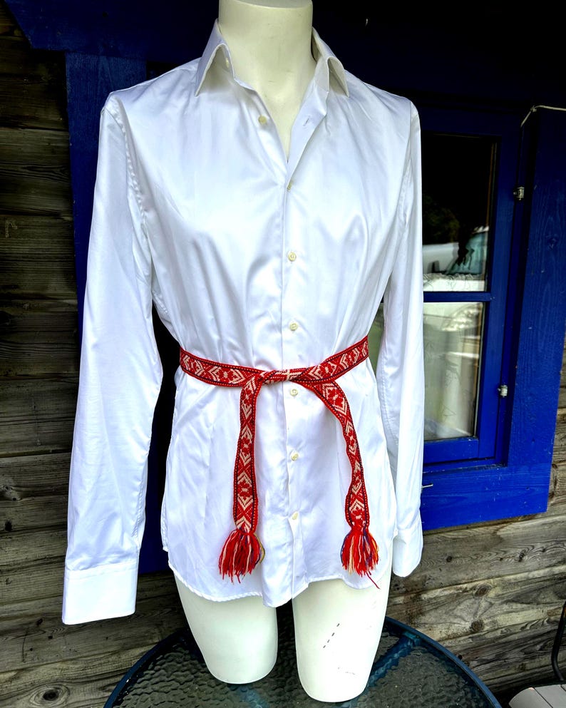 May include: A crisp white long-sleeved button-down shirt featuring a pointed collar and a central button placket. A narrow, woven belt with a red, white, and orange geometric pattern is tied around the waist, ending in two fringed tassels.