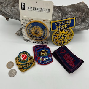 May include: A collection of embroidered patches in various shapes and sizes. The patches feature text and designs, including a sunburst, a cabin, and the words "STORLIEN" and "FJALL RAVEN". Some patches are blue and yellow, while others are red and black.