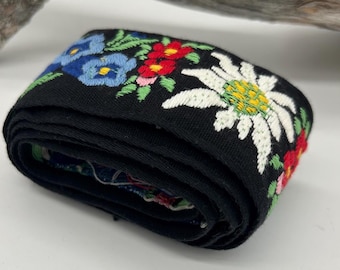 May include: A rolled-up black embroidered ribbon featuring colorful floral designs. The ribbon showcases blue, red, and white flowers with green leaves. The ribbon is likely used for decorative purposes.