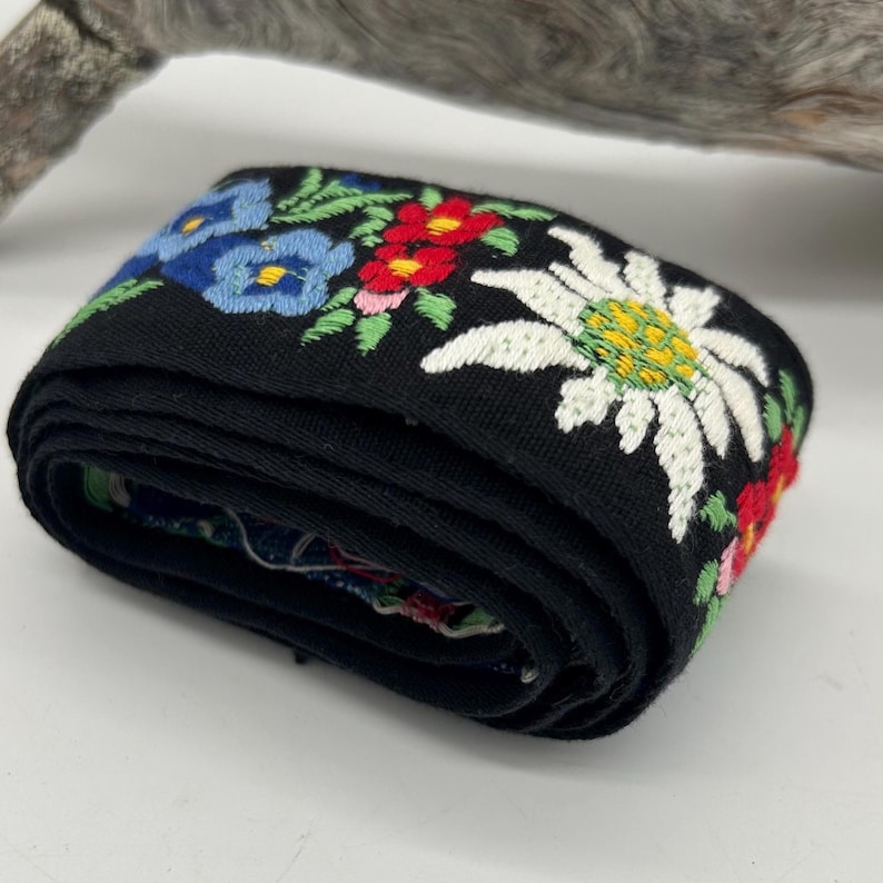 May include: A rolled black embroidered ribbon with colourful floral designs. The ribbon features blue, red, and white flowers with green leaves. The ribbon is likely used for decorative purposes.