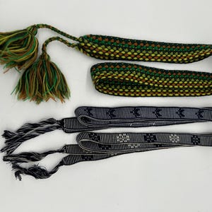 May include: An overhead shot shows three decorative textile items. The top item is a woven cord with green, yellow, and brown hues, and tassels. Below are two black and white patterned ribbons with tassels. The items are on a white background.