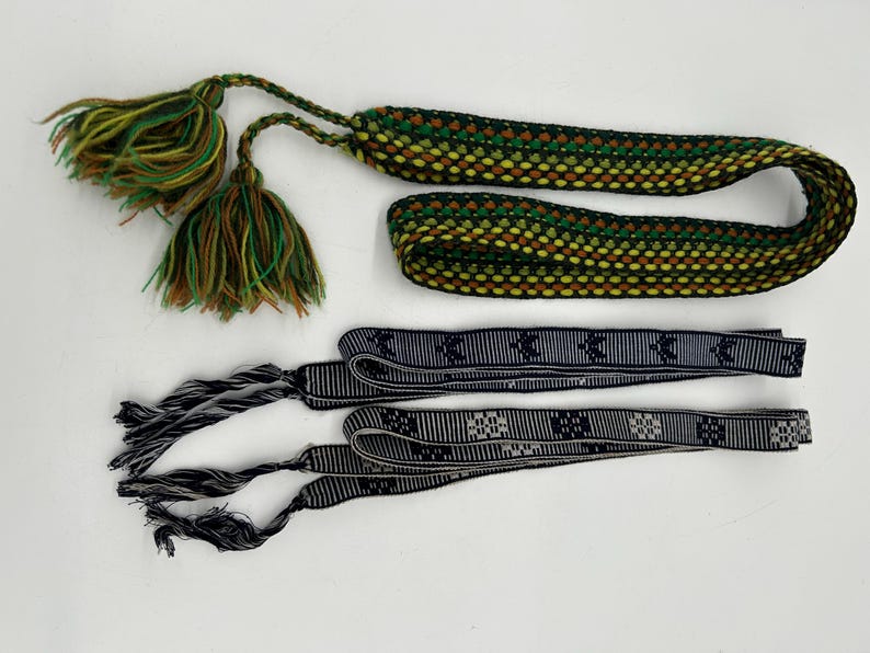 May include: An overhead shot shows three decorative textile items. The top item is a woven cord with green, yellow, and brown hues, and tassels. Below are two black and white patterned ribbons with tassels. The items are on a white background.