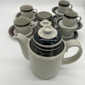 Puede incluir: Juego de té de cerámica vintage con tetera, tazas, platillos y azucarero. La tetera es de color gris claro con una tapa a rayas negras y marrones. Las tazas y platillos tienen un borde marrón.