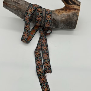 May include: Black ribbon with a repeating floral pattern in orange, yellow, and teal. The ribbon is draped over a piece of weathered wood, creating a rustic and decorative display.