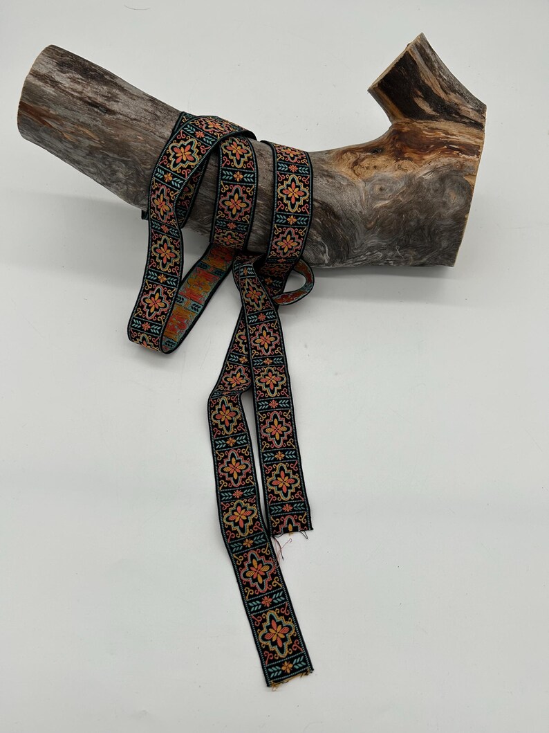 May include: Black ribbon with a repeating floral pattern in orange, yellow, and teal. The ribbon is draped over a piece of weathered wood, creating a rustic and decorative display.