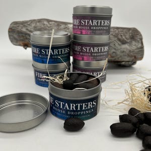 May include: A collection of small, silver tins containing fire starters, labelled "Fire Starters From Moose Droppings". The fire starters are dark brown and cylindrical. The tins are arranged on a white surface, with a piece of wood in the background.
