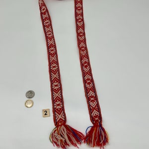 May include: A long, narrow woven belt featuring a repeating geometric pattern in red and white with thin blue border lines. The ends of the belt are finished with thick, multi-colored yarn tassels in red, white, blue, and yellow. A coin is visible for scale.