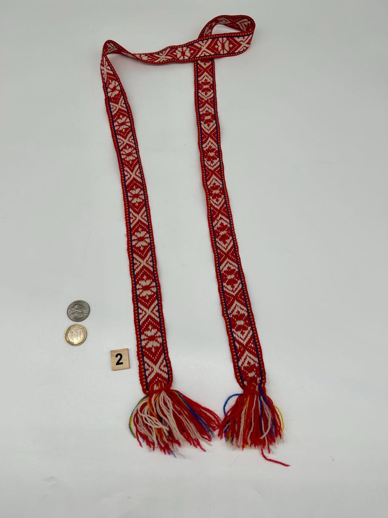 May include: A long, narrow woven belt featuring a repeating geometric pattern in red and white with thin blue border lines. The ends of the belt are finished with thick, multi-colored yarn tassels in red, white, blue, and yellow. A coin is visible for scale.