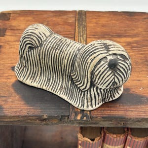 Può includere: Statuetta di cane in ceramica con un motivo a strisce crema e nere. Il cane è sdraiato, con la testa e il corpo dettagliati con linee verticali. La statuetta è posta su una mensola di legno.