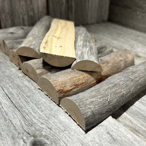 May include: A collection of cut firewood logs, with a mix of gray and light brown wood tones. The logs are arranged on a weathered wooden surface, creating a rustic and natural aesthetic. The wood has visible grain and texture.