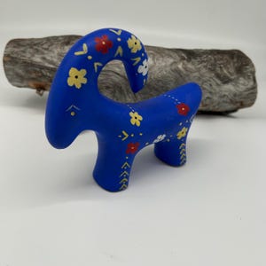 May include: A blue ceramic goat figurine with yellow, red, and white floral accents. The goat has a curved neck and is standing on four legs. The figurine is placed in front of a piece of driftwood.