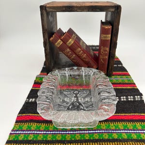 Peut inclure: Un cendrier carré en verre transparent avec un motif de plumes détaillé. Le cendrier est posé sur un textile coloré à motifs. Derrière, une caisse en bois contient plusieurs livres anciens avec des couvertures rouges et des lettres dorées.