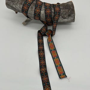 May include: Black ribbon with a repeating floral pattern in orange, teal, and yellow. The ribbon is wrapped around a piece of driftwood. The ribbon is approximately 1 inch wide.