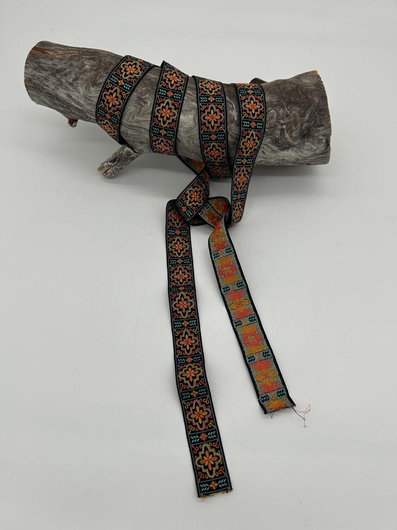 May include: Black ribbon with a repeating floral pattern in orange, teal, and yellow. The ribbon is wrapped around a piece of driftwood. The ribbon is approximately 1 inch wide.
