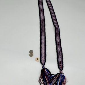 May include: A long, woven textile belt or strap featuring a repeating striped pattern in black, white, red, and blue. The ends of the strap are finished with thick, multi-colored yarn tassels in matching shades. A small square tile with the number 3 is visible.