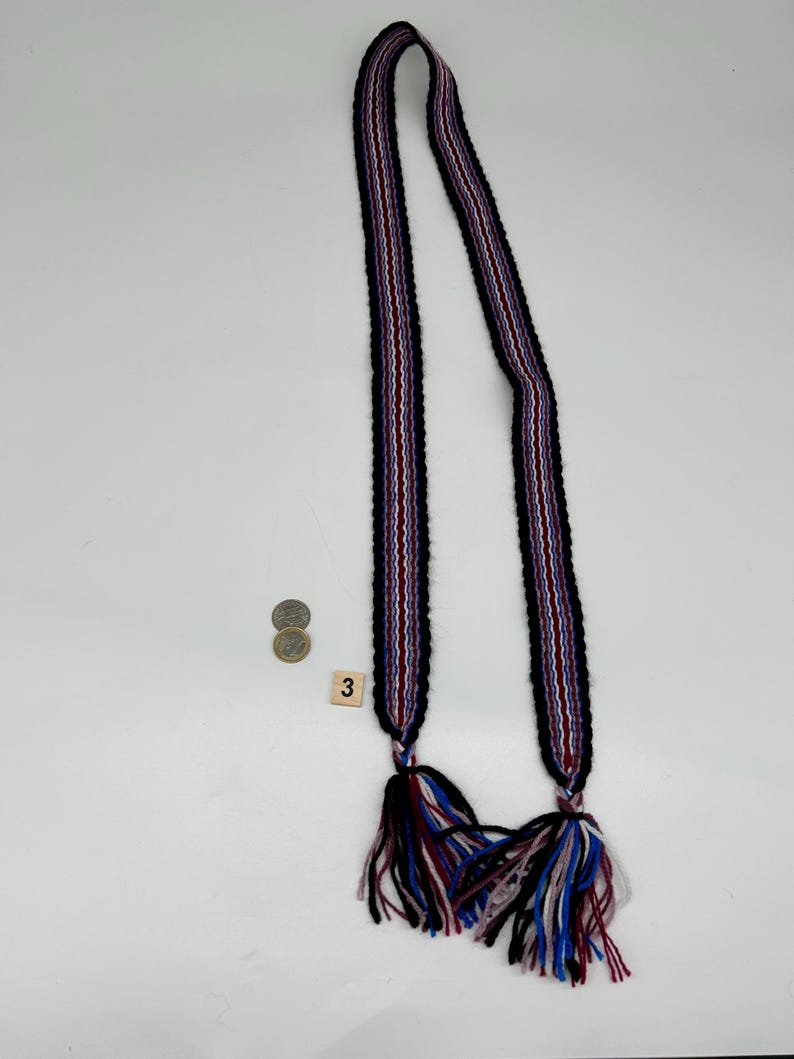 May include: A long, woven textile belt or strap featuring a repeating striped pattern in black, white, red, and blue. The ends of the strap are finished with thick, multi-colored yarn tassels in matching shades. A small square tile with the number 3 is visible.